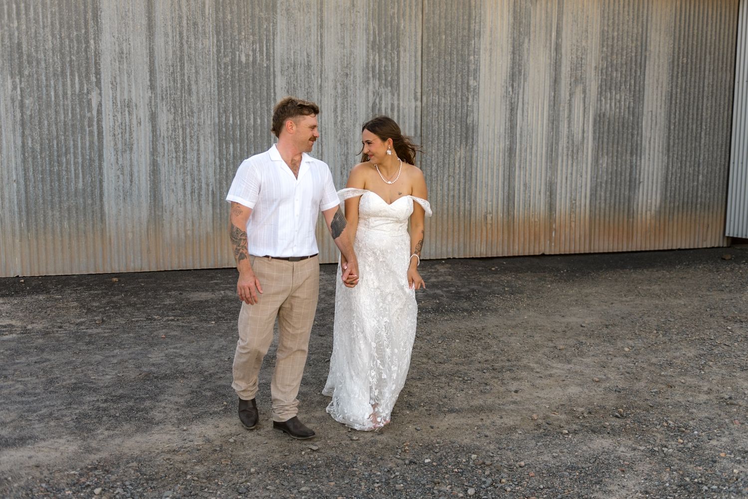 Smiling bride and groom walk hand-in-hand in front of a large corrugated metal wall on their wedding day.