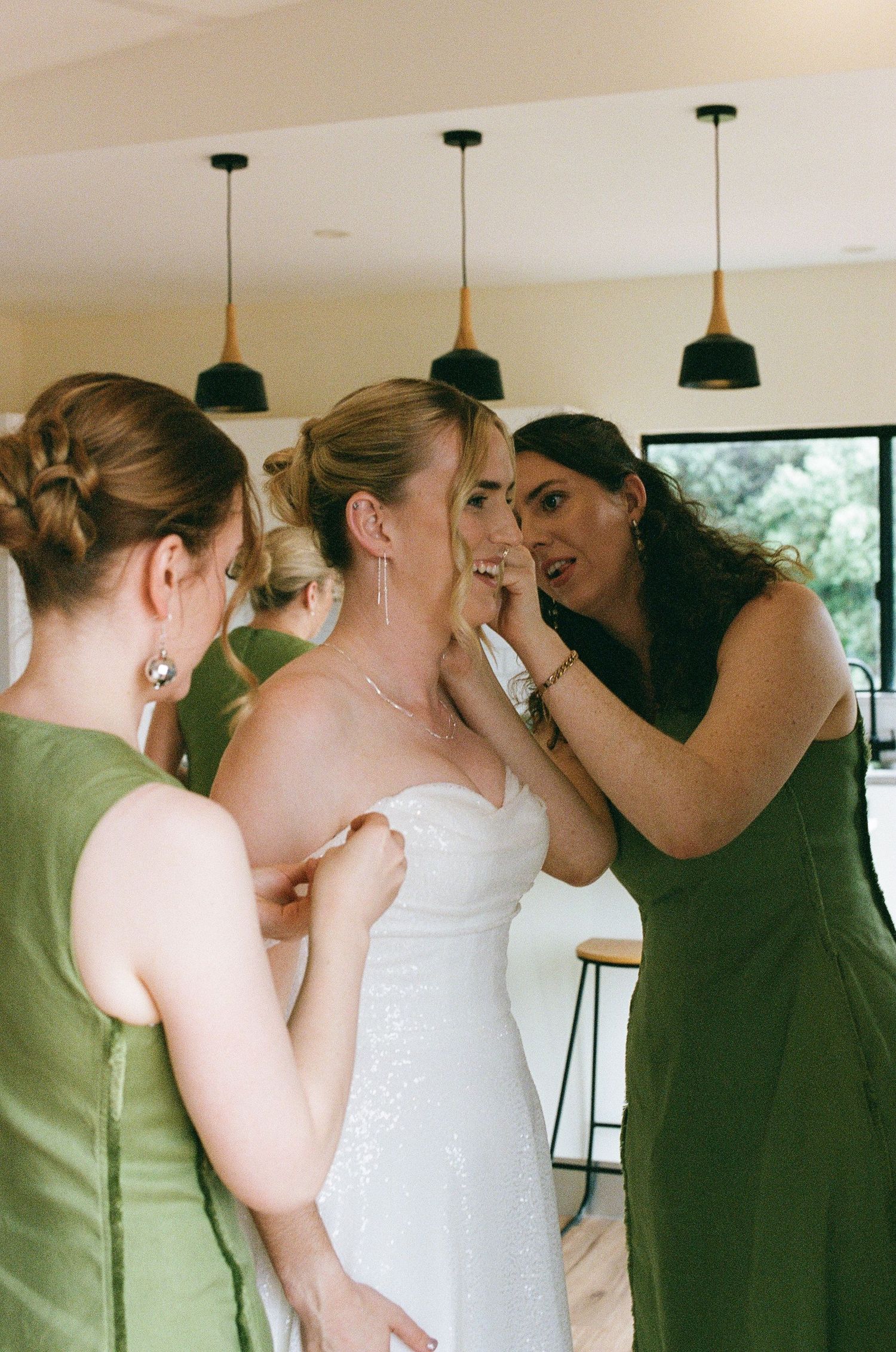 Bridesmaids in green dresses help bride with jewelry and dress details before wedding ceremony.