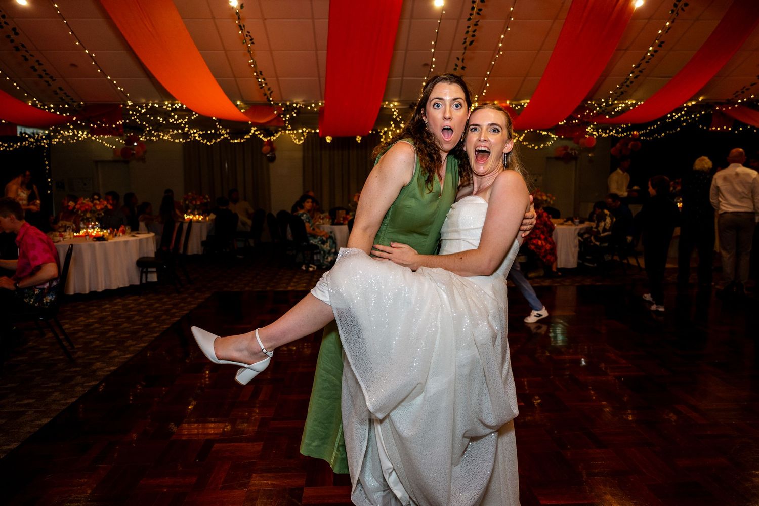 Two women celebrate at a wedding reception, one in green lifting the bride in white amid festive lights.