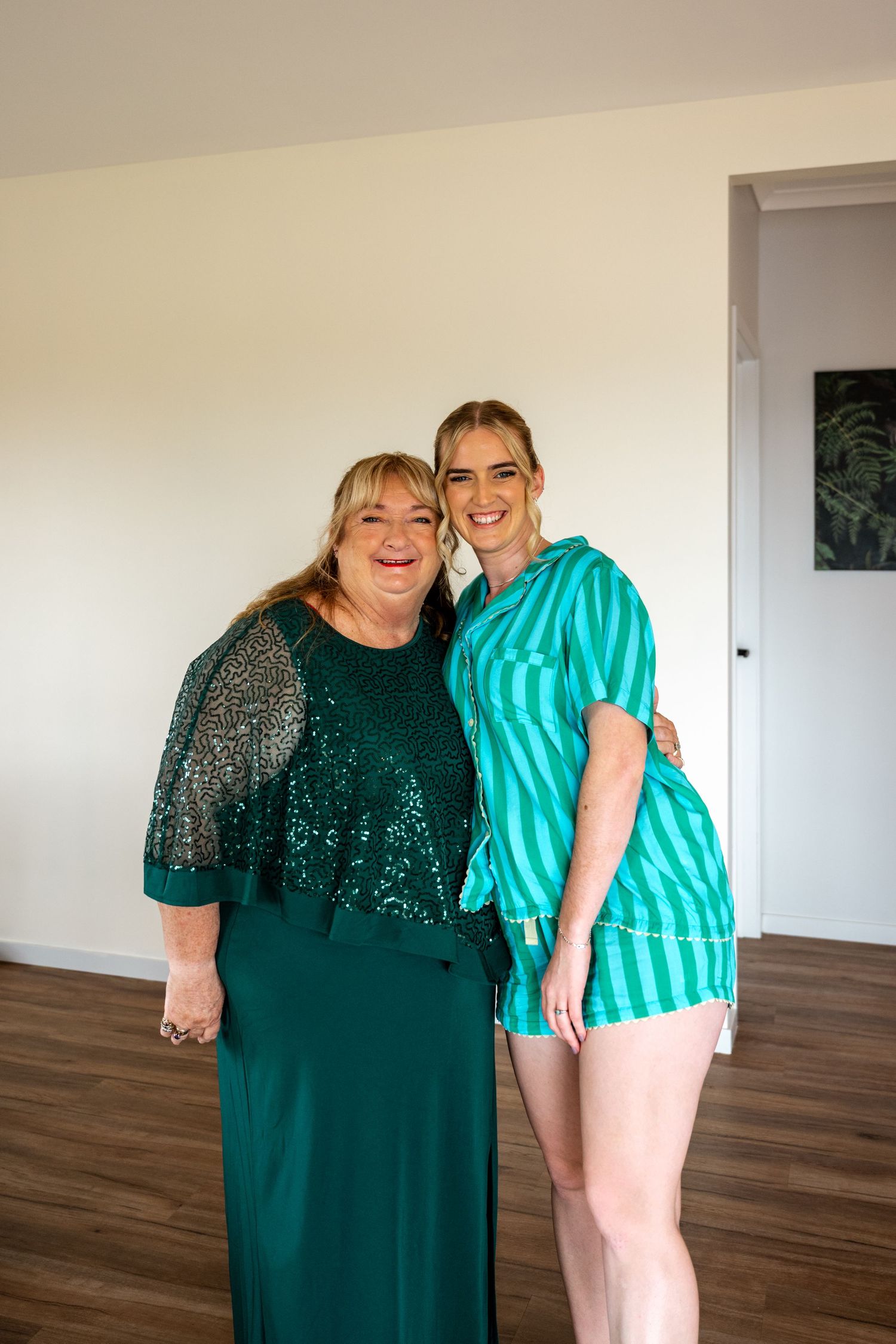 Two women in matching teal outfits smiling together during bridal preparations in bright room.