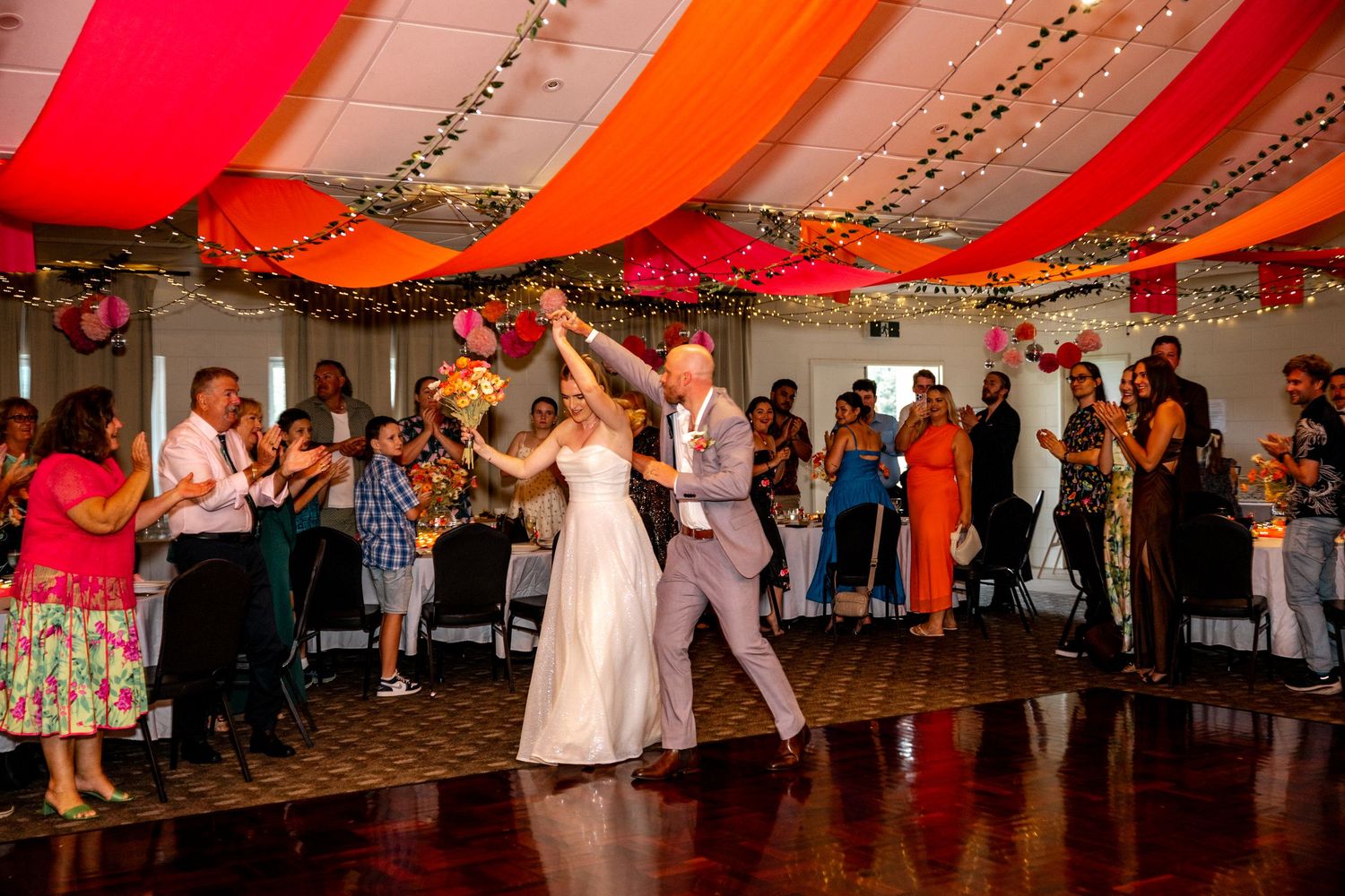 Bride and groom sharing their first dance under vibrant orange and red draped ceiling with fairy lights at wedding reception.