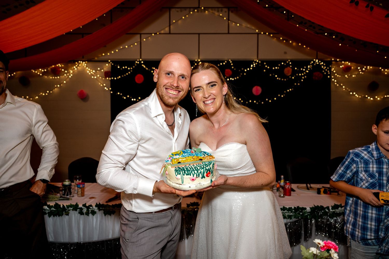 Smiling bride and groom proudly hold a colorful decorated wedding cake.