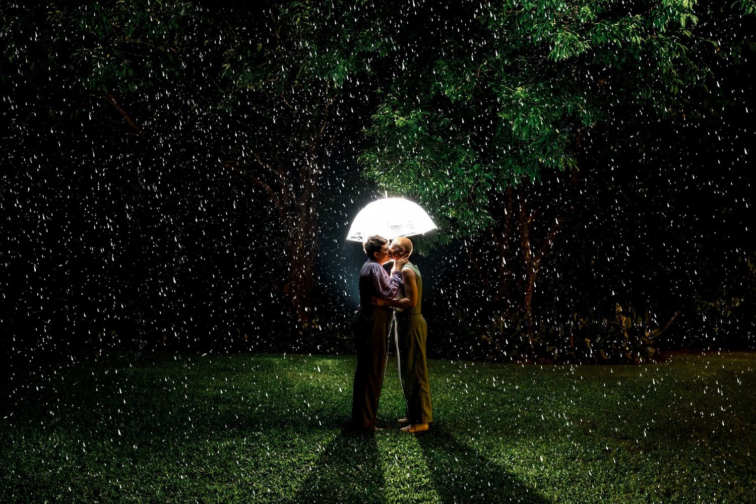 Couple kissing under a glowing umbrella in the rain at night outdoors.