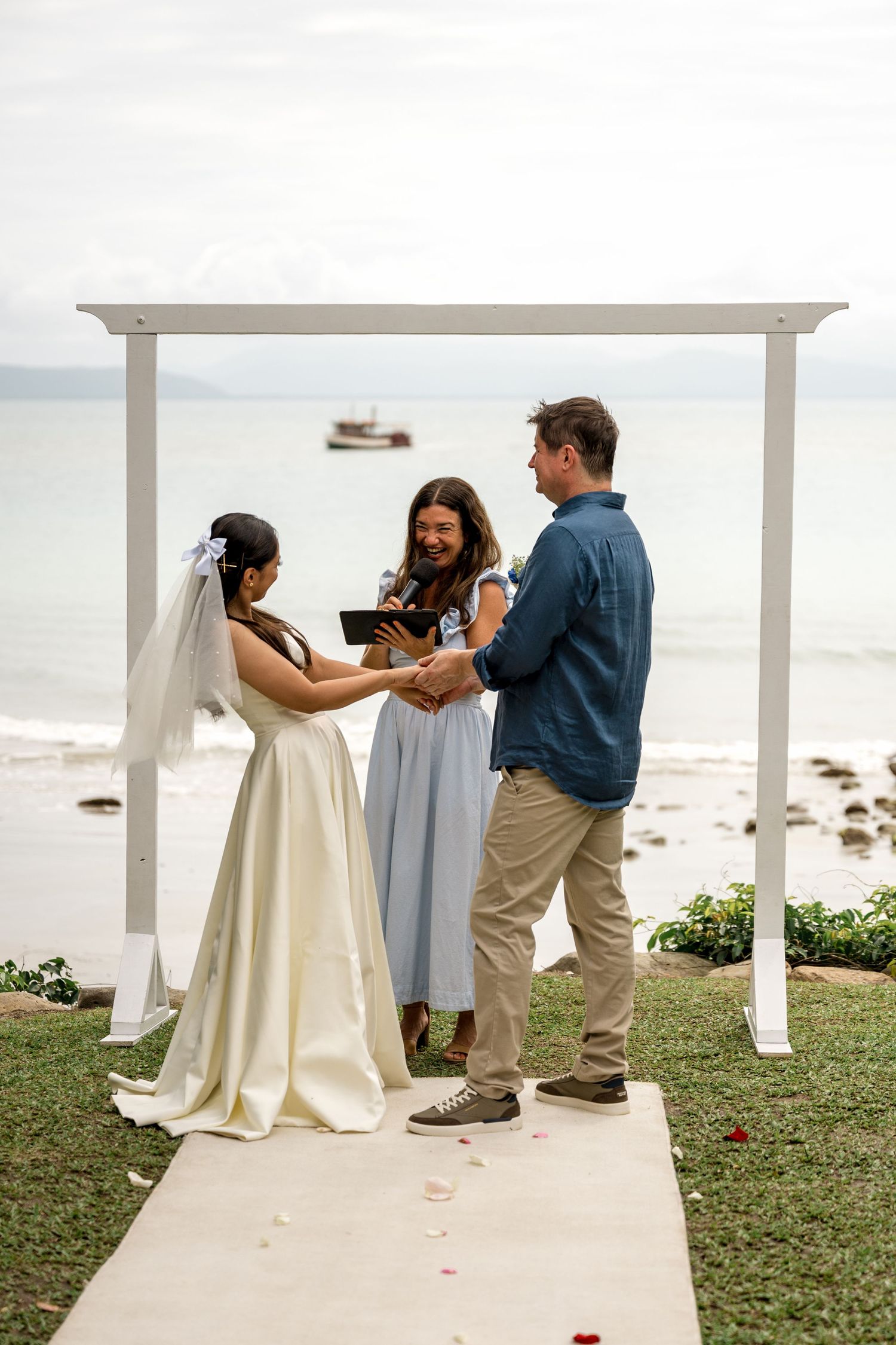Bride reads vows to groom under a white arch by the ocean, officiated by a woman in a blue dress at Little Cove, Port Douglas.