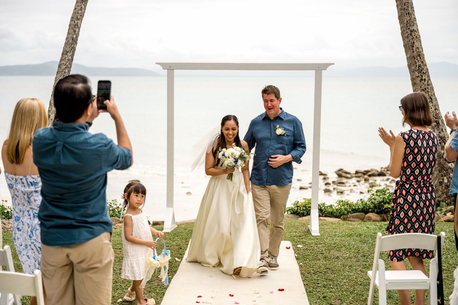 Bride walks down aisle with groom at tropical beach wedding ceremony with white arch and palm trees at Little Cove, Port Douglas.