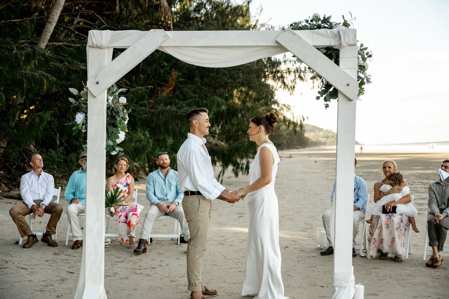 Bride and groom holding hands at an outdoor beach wedding ceremony with seated guests watching under a rustic white arch at Four Mile Beach.