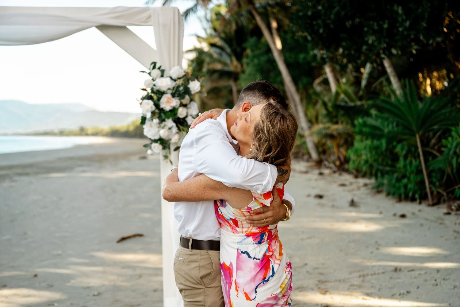 Groom embracing a woman in a colorful dress during a beach wedding celebration under a floral arch at Four Mile Beach, Port Douglas.