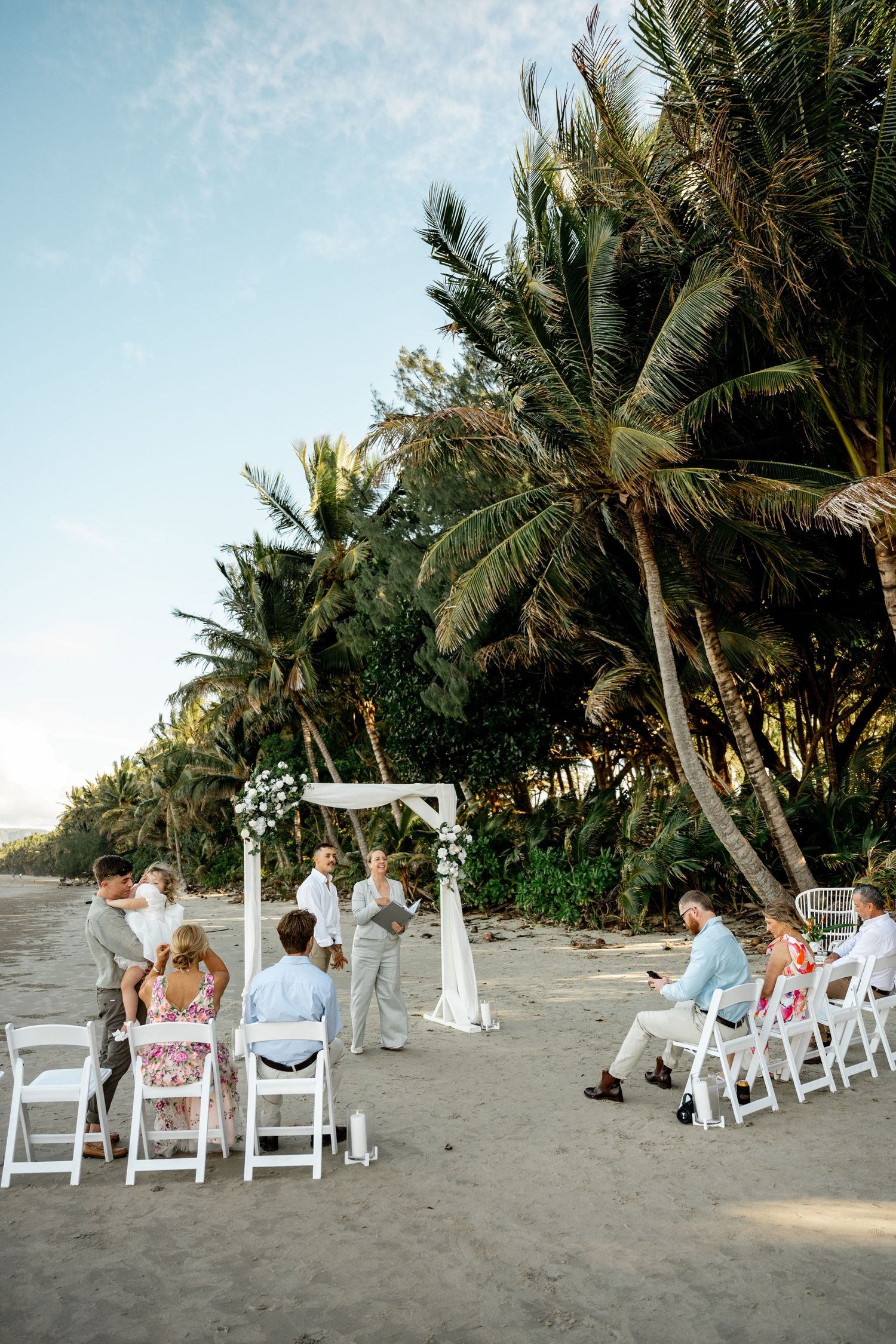 Small intimate beach wedding ceremony with guests seated in white chairs under palm trees at sunset at Four Mile Beach, Port Douglas.