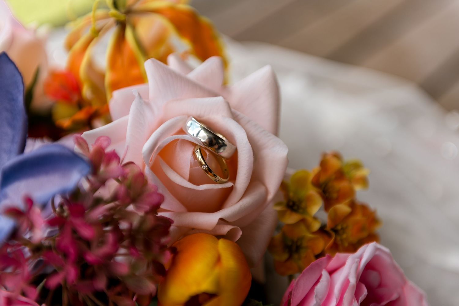 Gold wedding rings resting inside a pink rose bloom surrounded by vibrant orange and pink bouquet flowers.