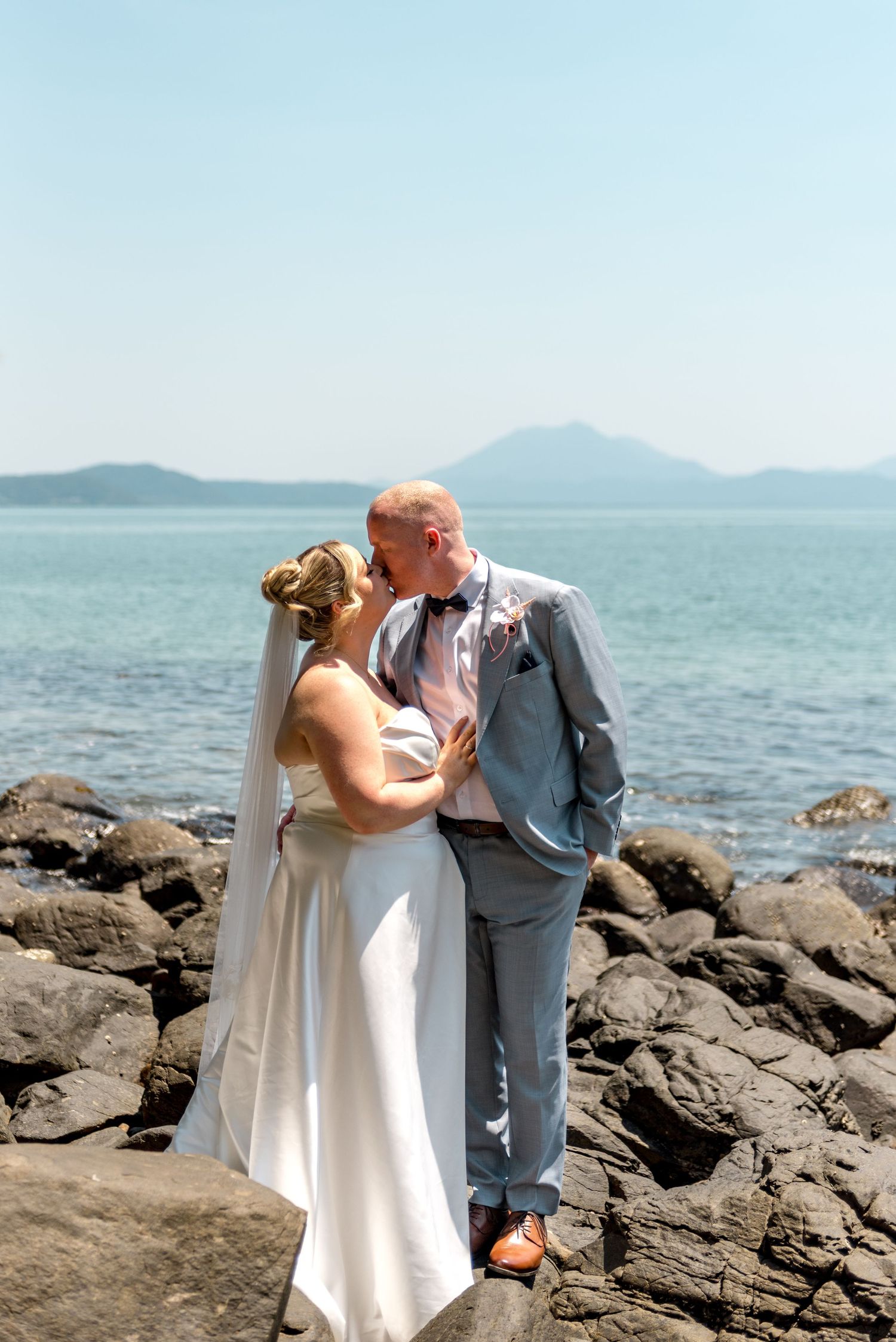 Bride and groom share a tender kiss on rocky beach with calm turquoise sea and mountains behind.