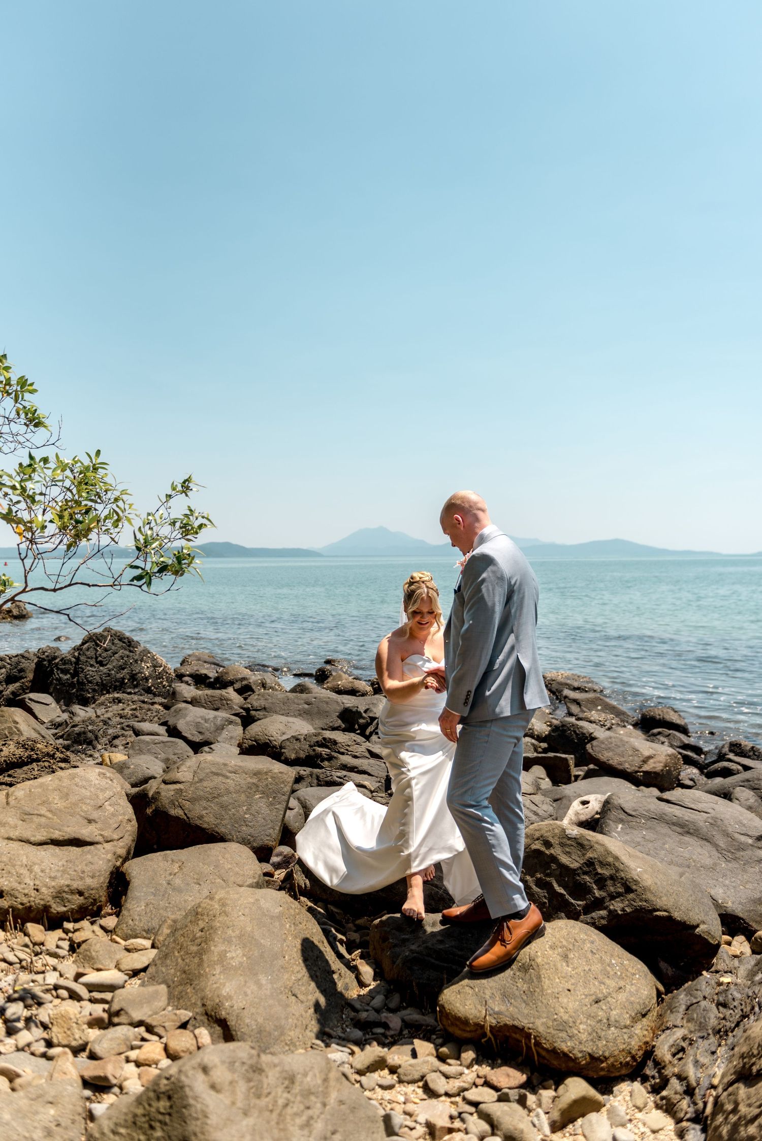 Bride and groom navigate rocky coastal terrain hand-in-hand with ocean and mountain views behind them.
