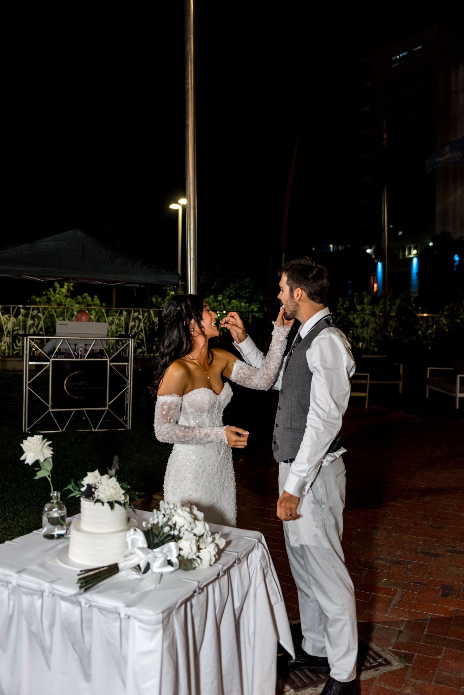 Bride and groom share wedding cake cutting moment outdoors at night.