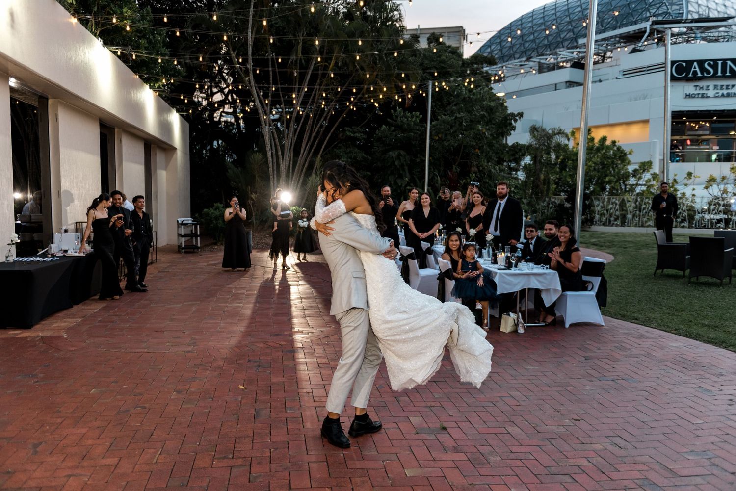 Groom lifting bride during romantic first dance on brick patio at outdoor evening wedding reception with guests watching.