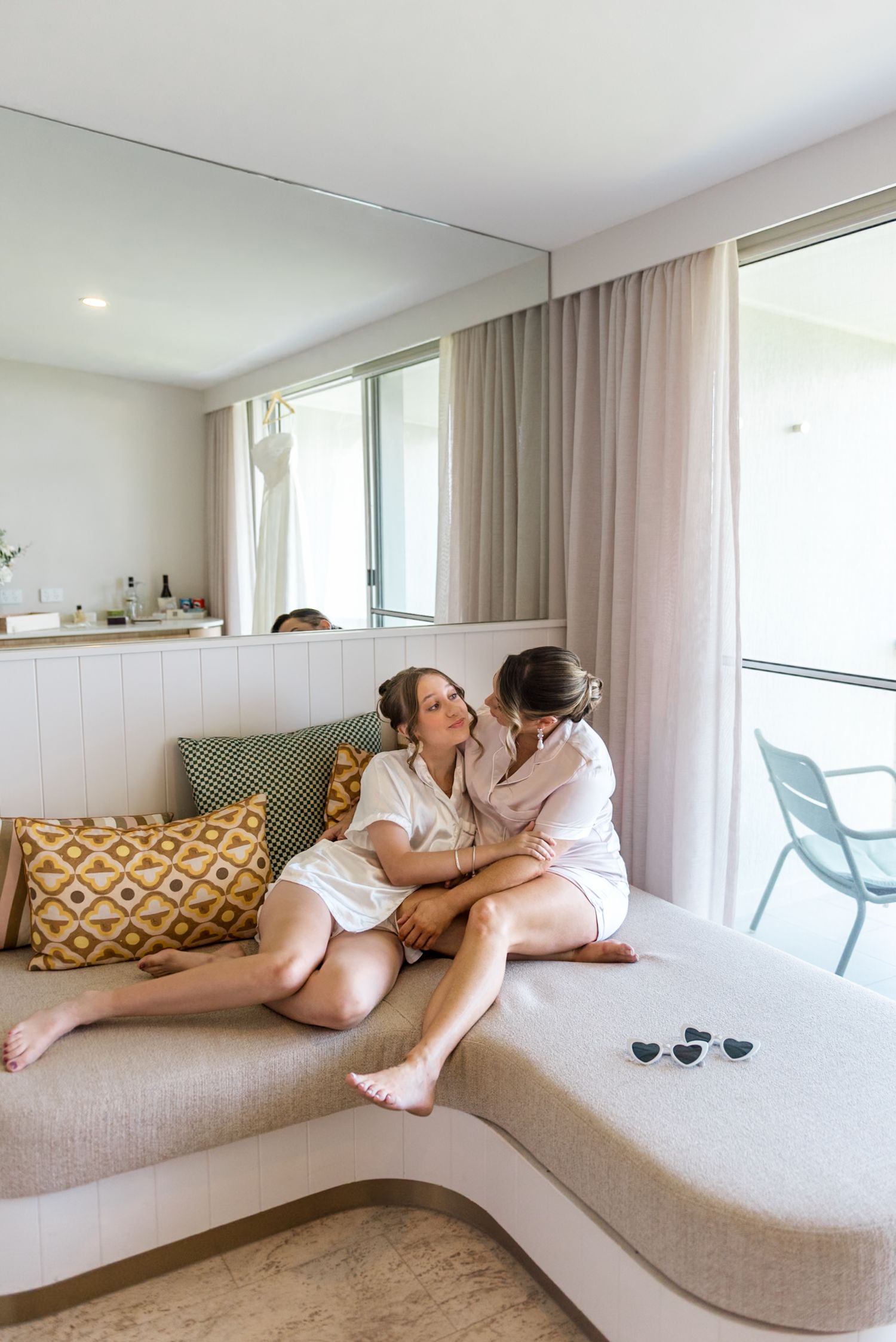 Two women in white robes sitting closely together on a beige sofa in a bright hotel room.