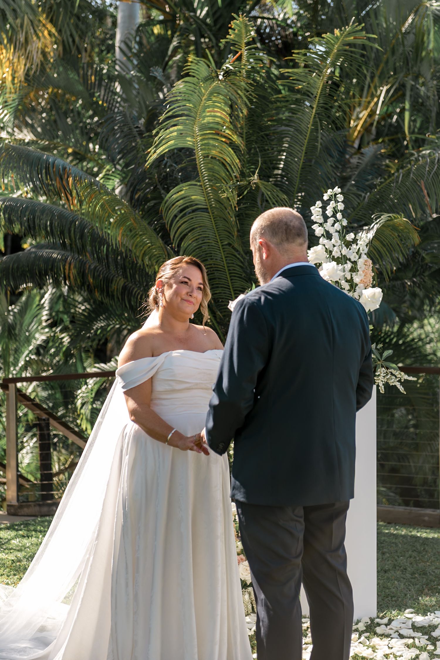 Bride in off-shoulder white gown holds hands with groom at outdoor wedding altar surrounded by tropical foliage.
