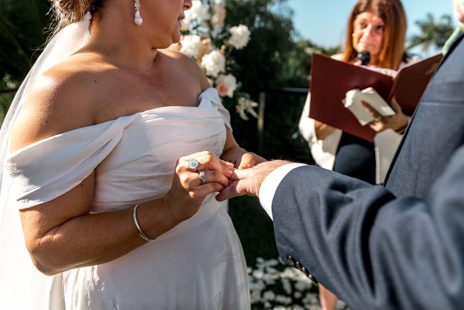 Bride placing wedding ring on groom's finger during outdoor ceremony with white floral arch.