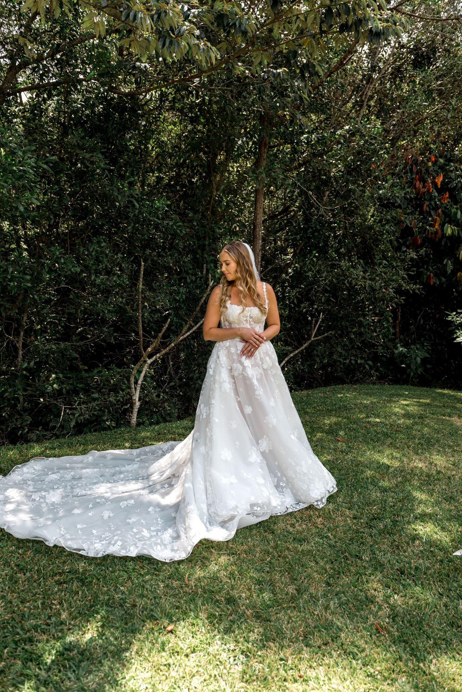 Bride in a stunning floral applique A-line wedding gown with long train standing in a sunlit garden.