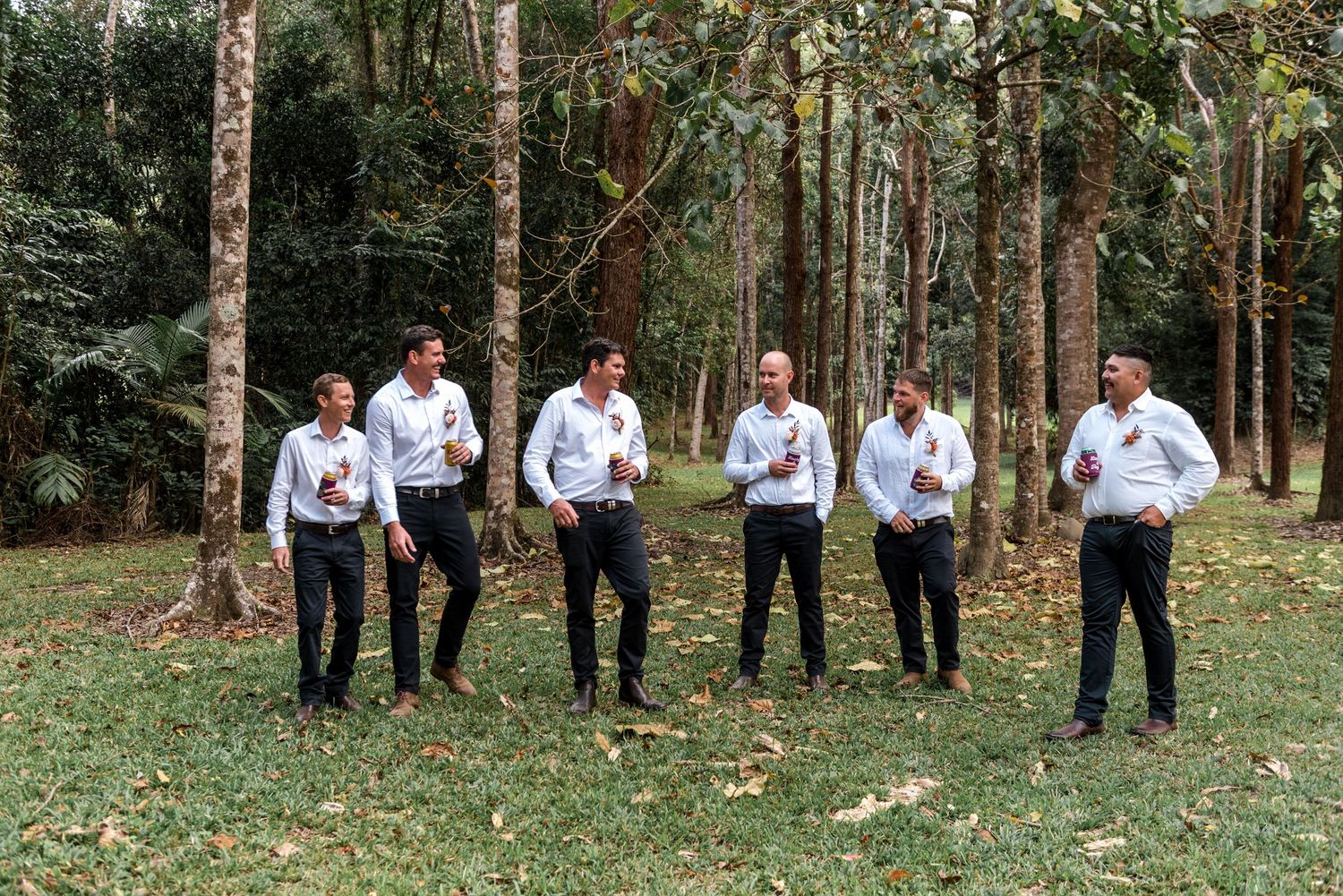 Six groomsmen in white shirts and dark pants stand on grass among tall trees at an outdoor wedding.