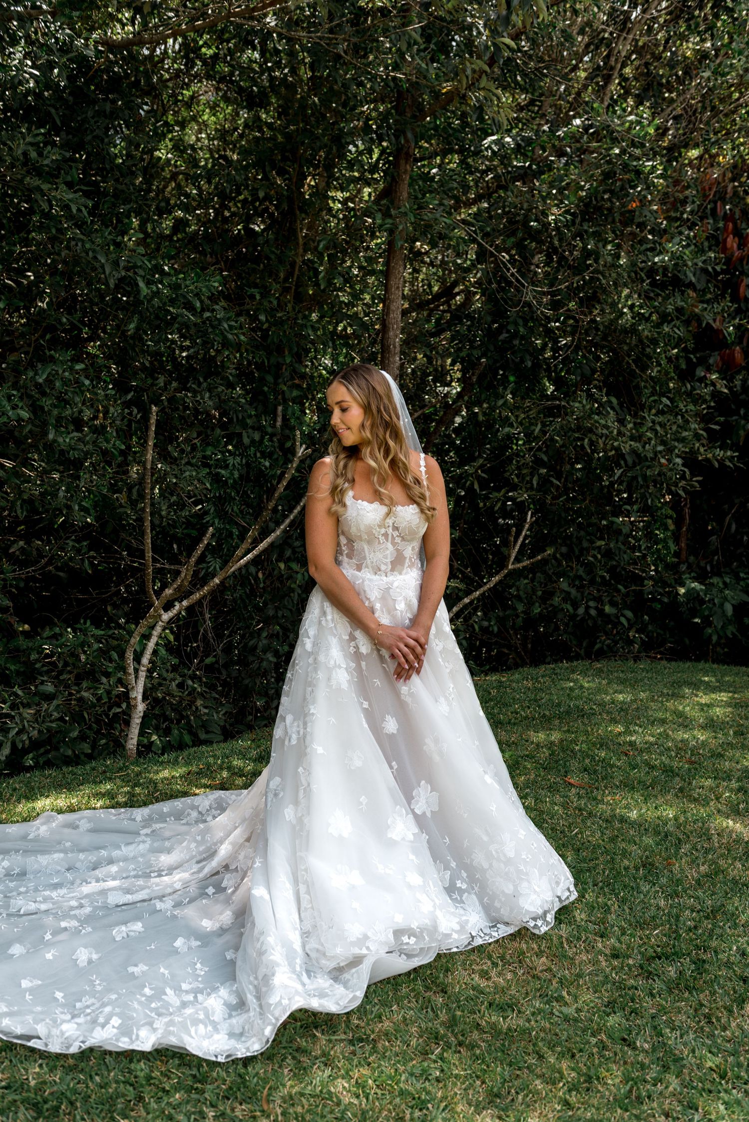 Beautiful bride in a floral lace wedding dress with cathedral train posing among green trees outdoors.