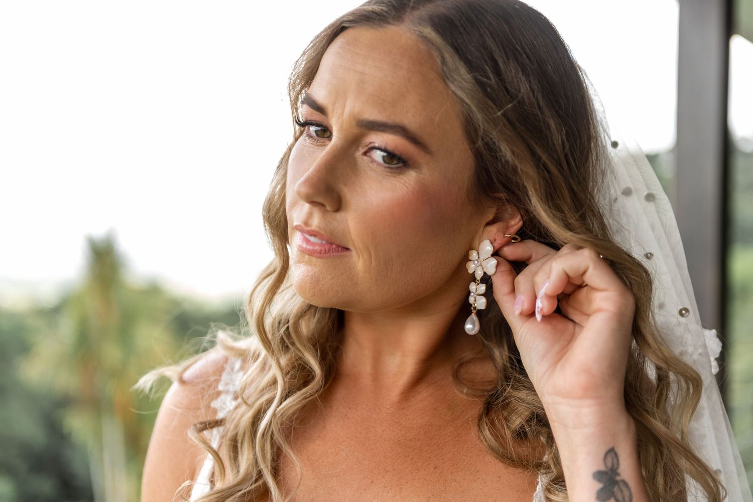 Bride adjusts pearl drop earring while wearing veil and lace gown on wedding day.