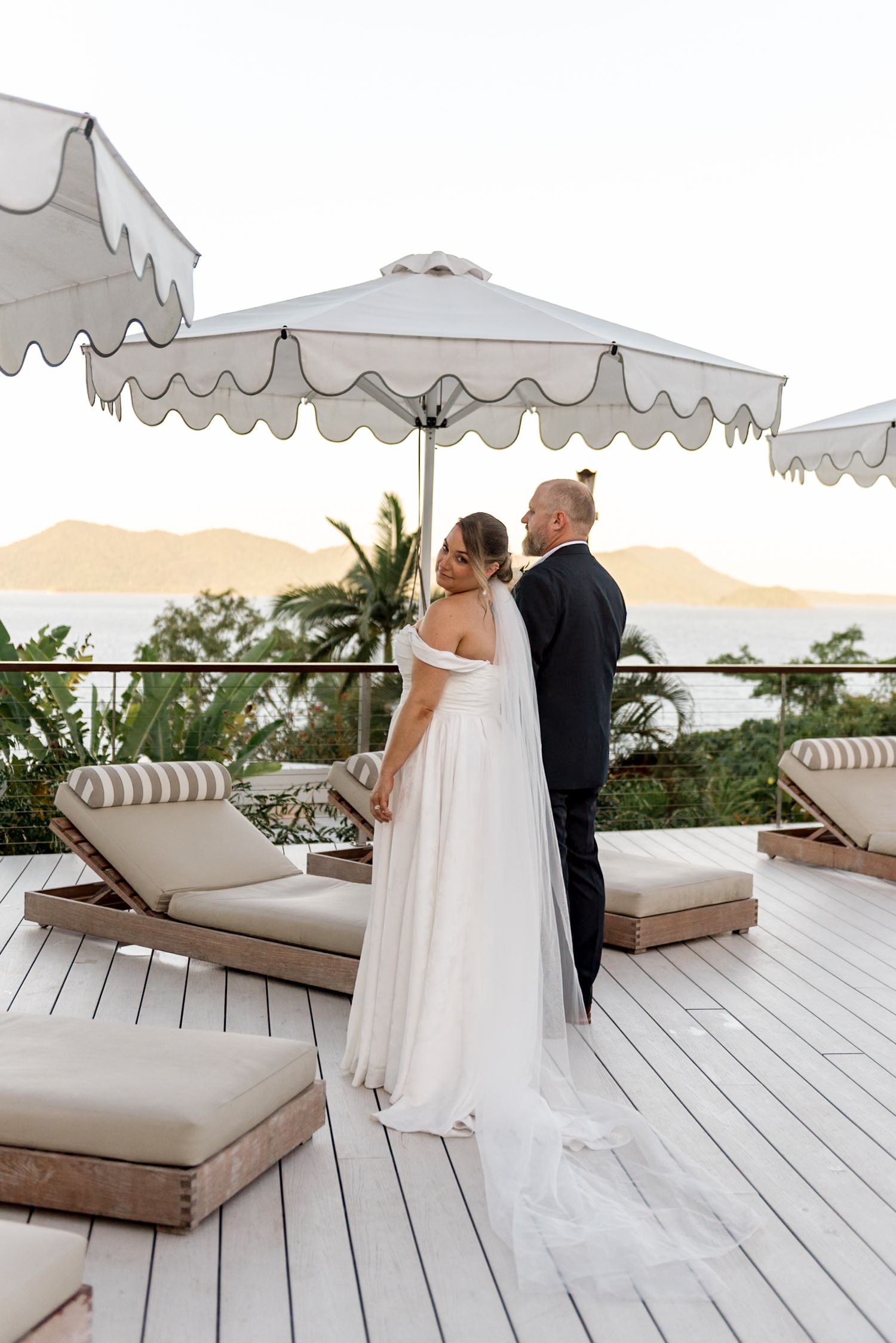 Bride and groom embracing under a white scalloped umbrella on a deck with ocean views.