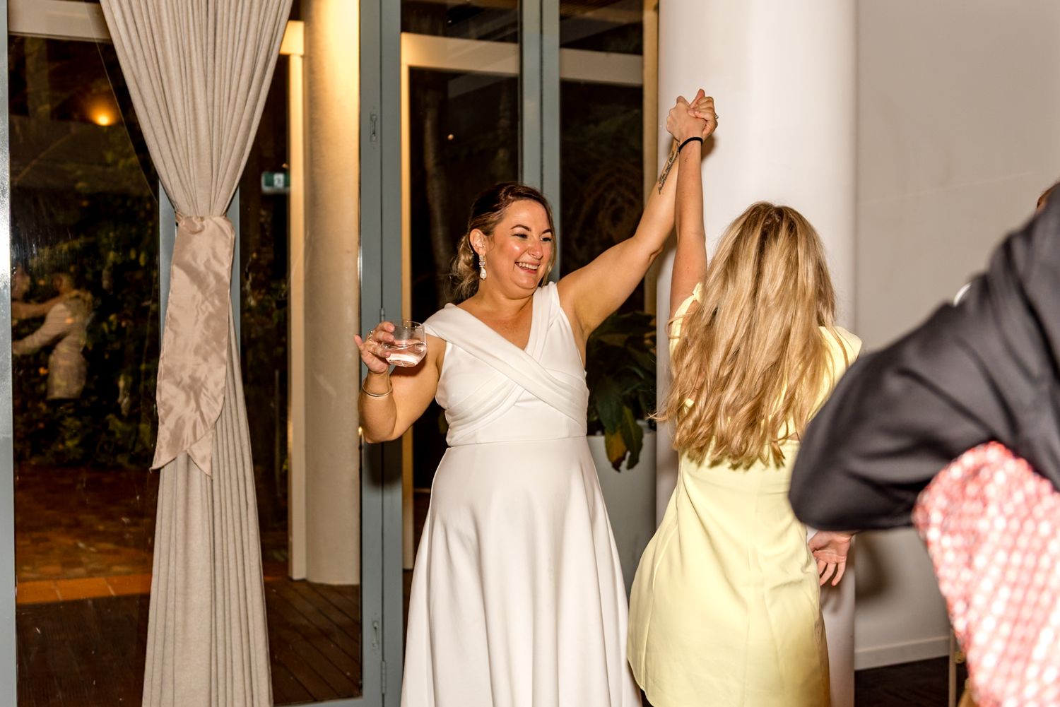 Bride in white dress dances joyfully with a guest in a yellow dress.