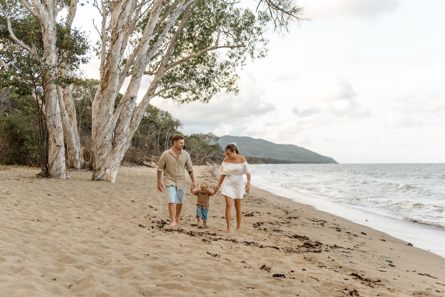 Family of three walking hand in hand along sandy beach with white gum trees and ocean.