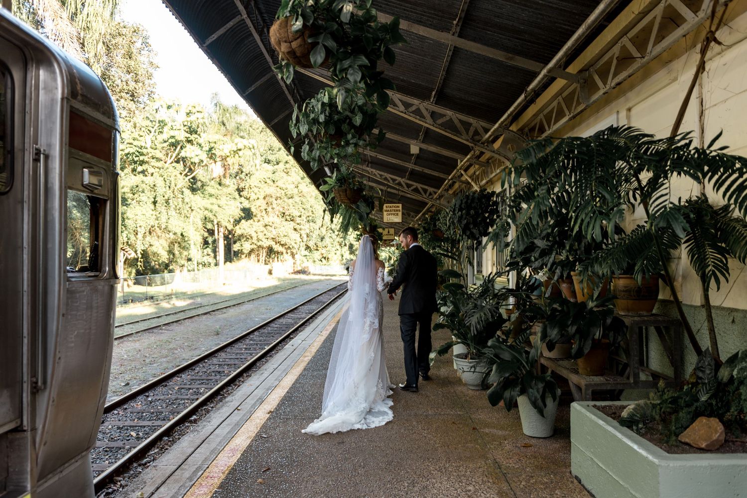 Bride in flowing white veil stands with groom on a vintage train station platform beside the tracks.