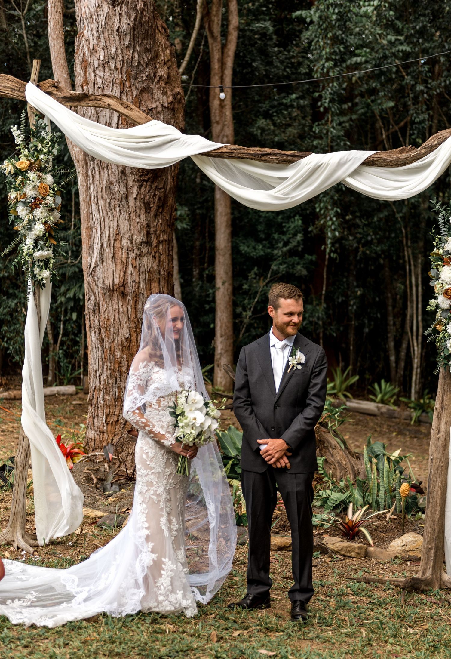 Bride in lace gown and veil standing with groom at rustic woodland wedding ceremony arch.