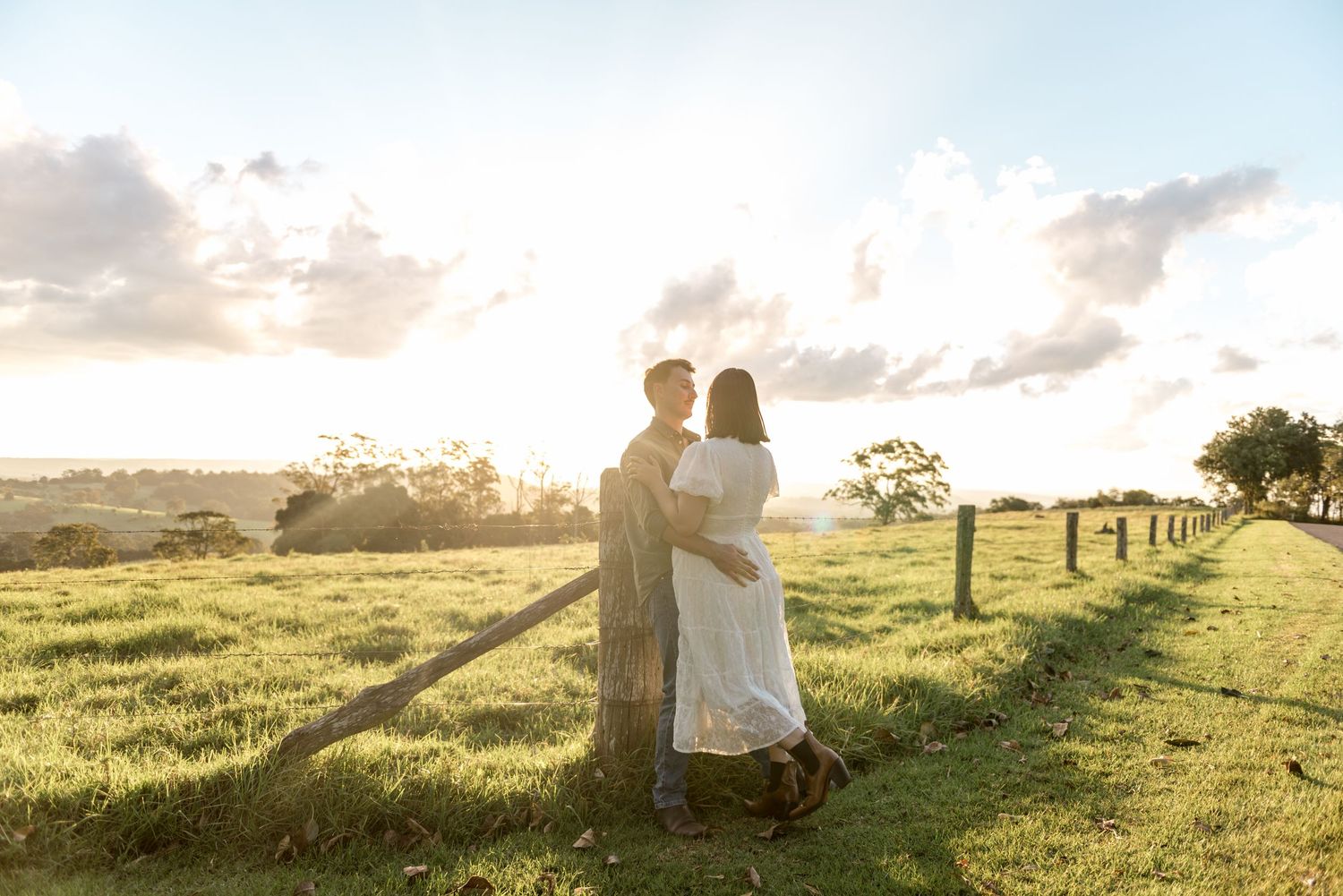 Couple embracing by a rustic fence at sunset overlooking a vast green landscape.
