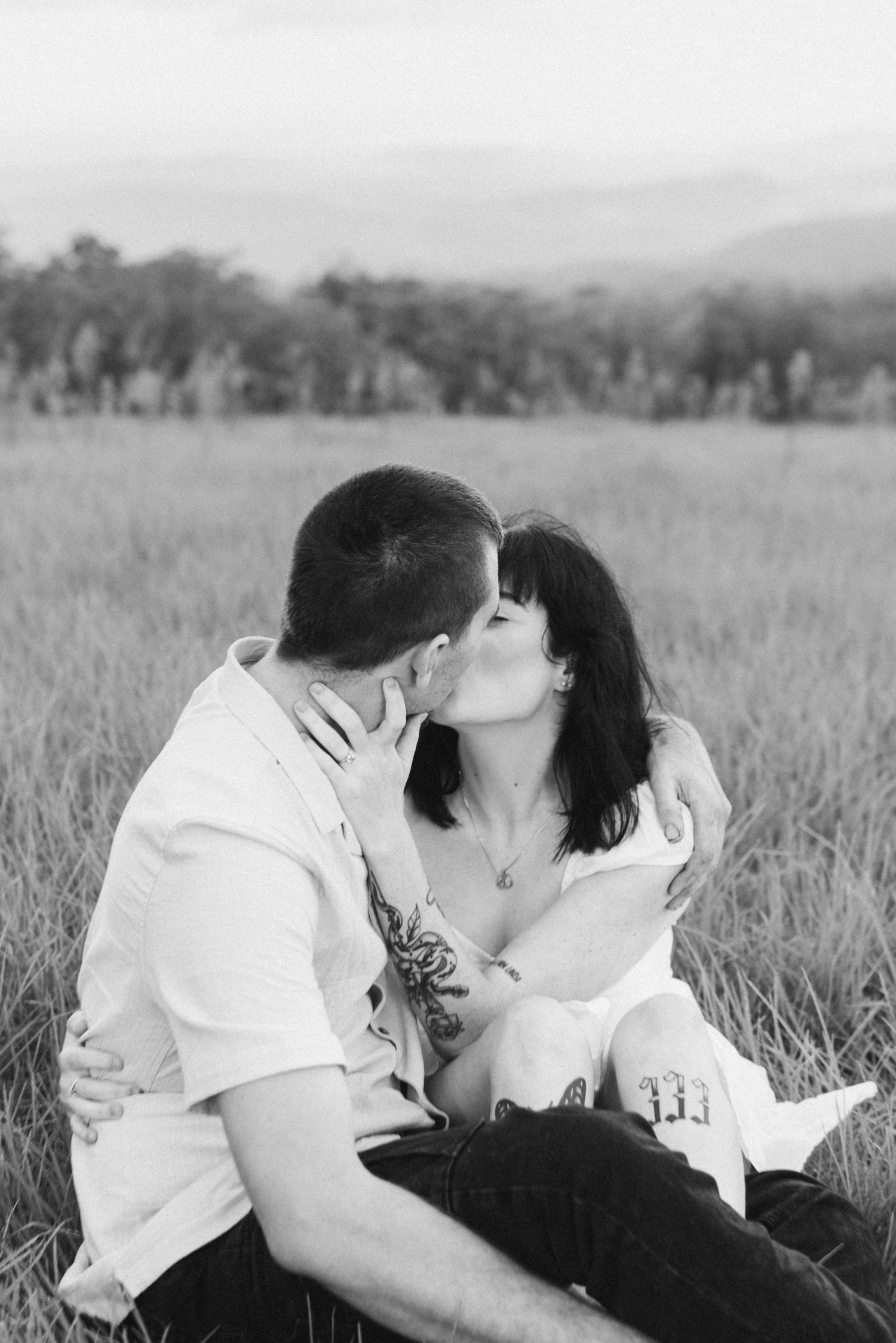 Black and white romantic photo of couple kissing in open field with mountain backdrop.