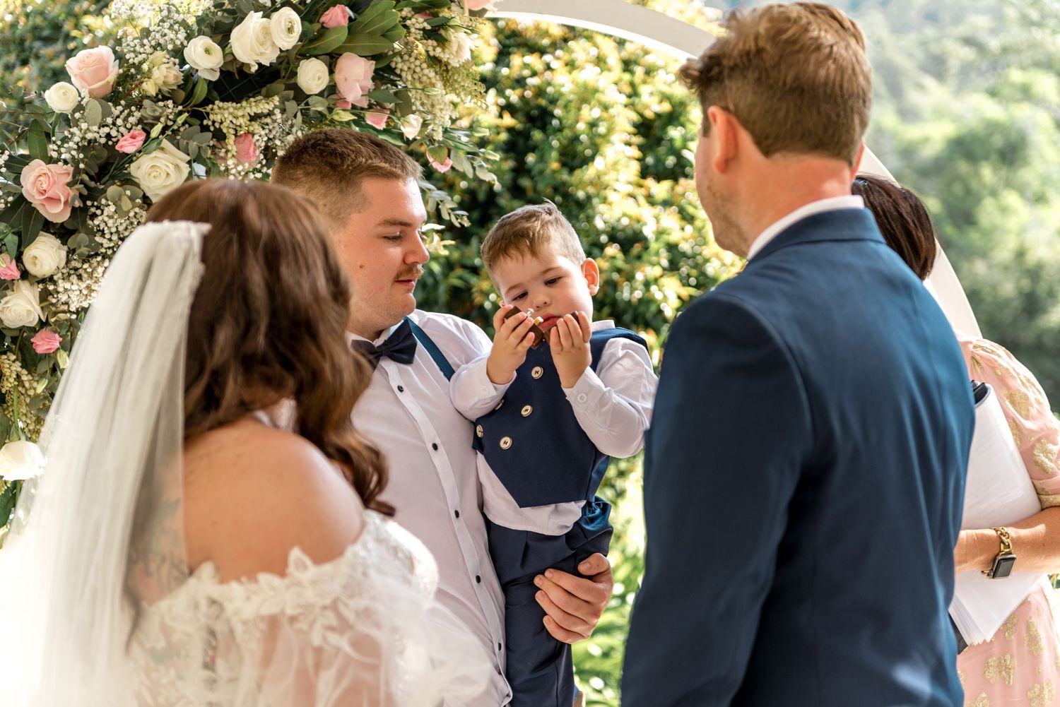 Young boy in navy suit laughing during outdoor wedding ceremony beside bride and groom at floral arch.