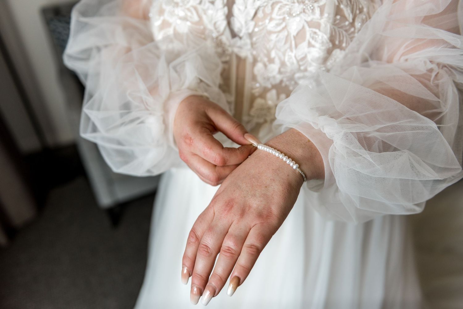 Bride in lace tulle gown fastening pearl bracelet on wrist with manicured nails.