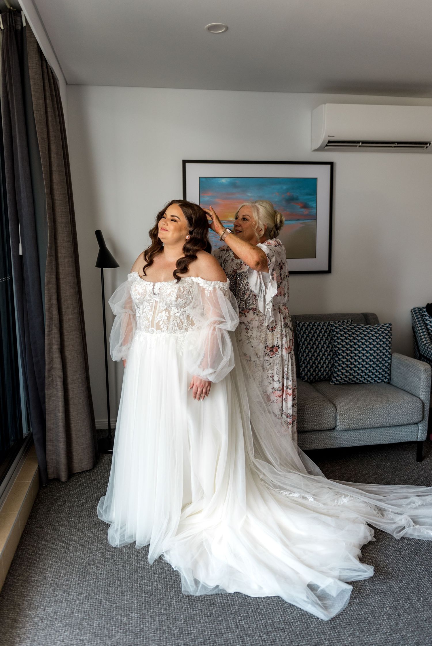 Bride in flowing tulle wedding gown having veil adjusted by woman in hotel room.