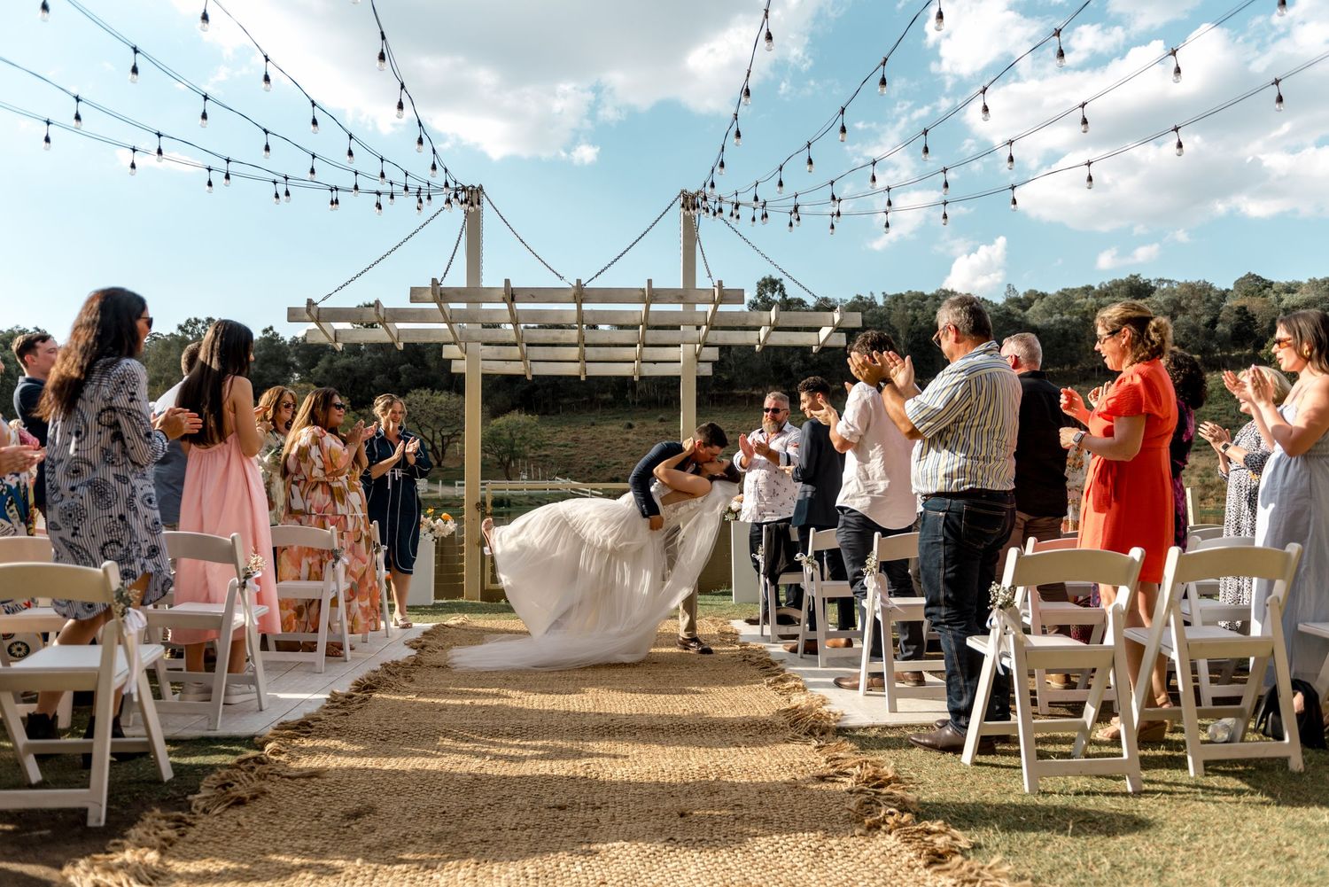 Bride and groom dip kiss during recessional at outdoor wedding with cheering guests and string lights above.