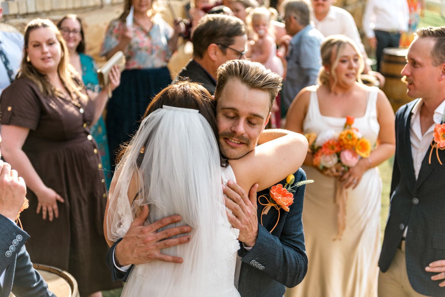 Groom embraces a guest during outdoor wedding reception with bridesmaids holding orange bouquets in background.