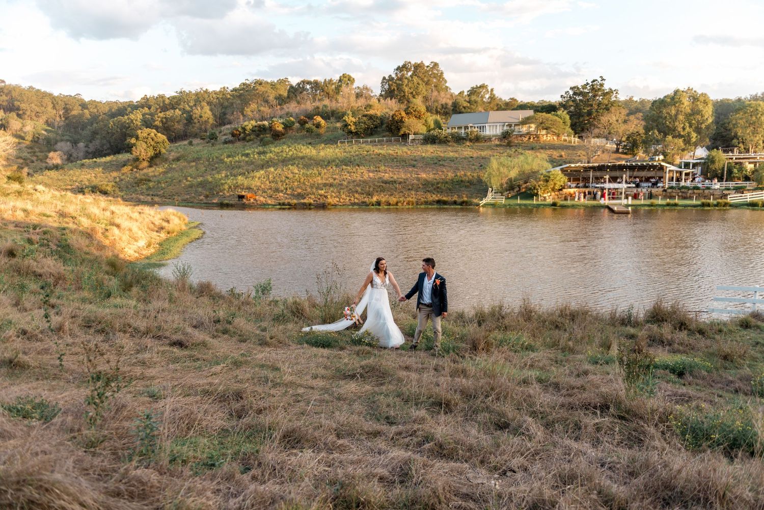 Bride and groom walk hand in hand along a grassy riverbank with a scenic waterway and venue behind them.