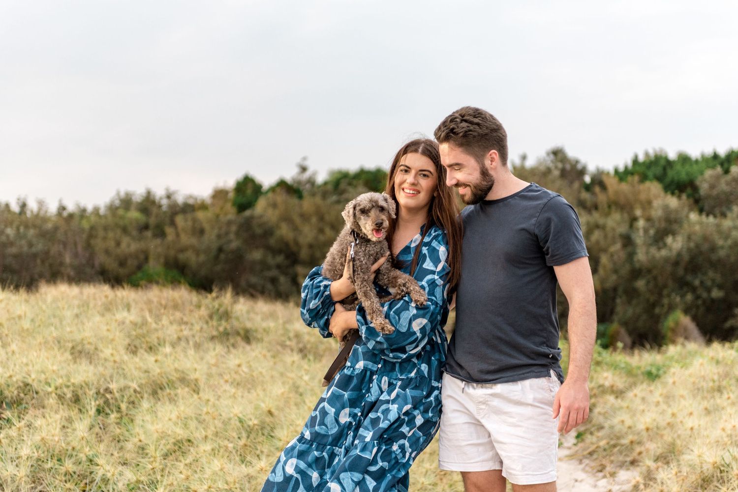 Smiling couple holding grey dog in open grassy coastal landscape.
