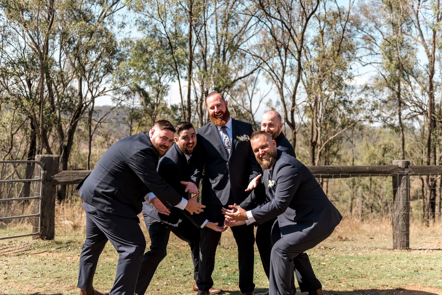 Five groomsmen in navy suits posing playfully together at outdoor rustic wedding venue.