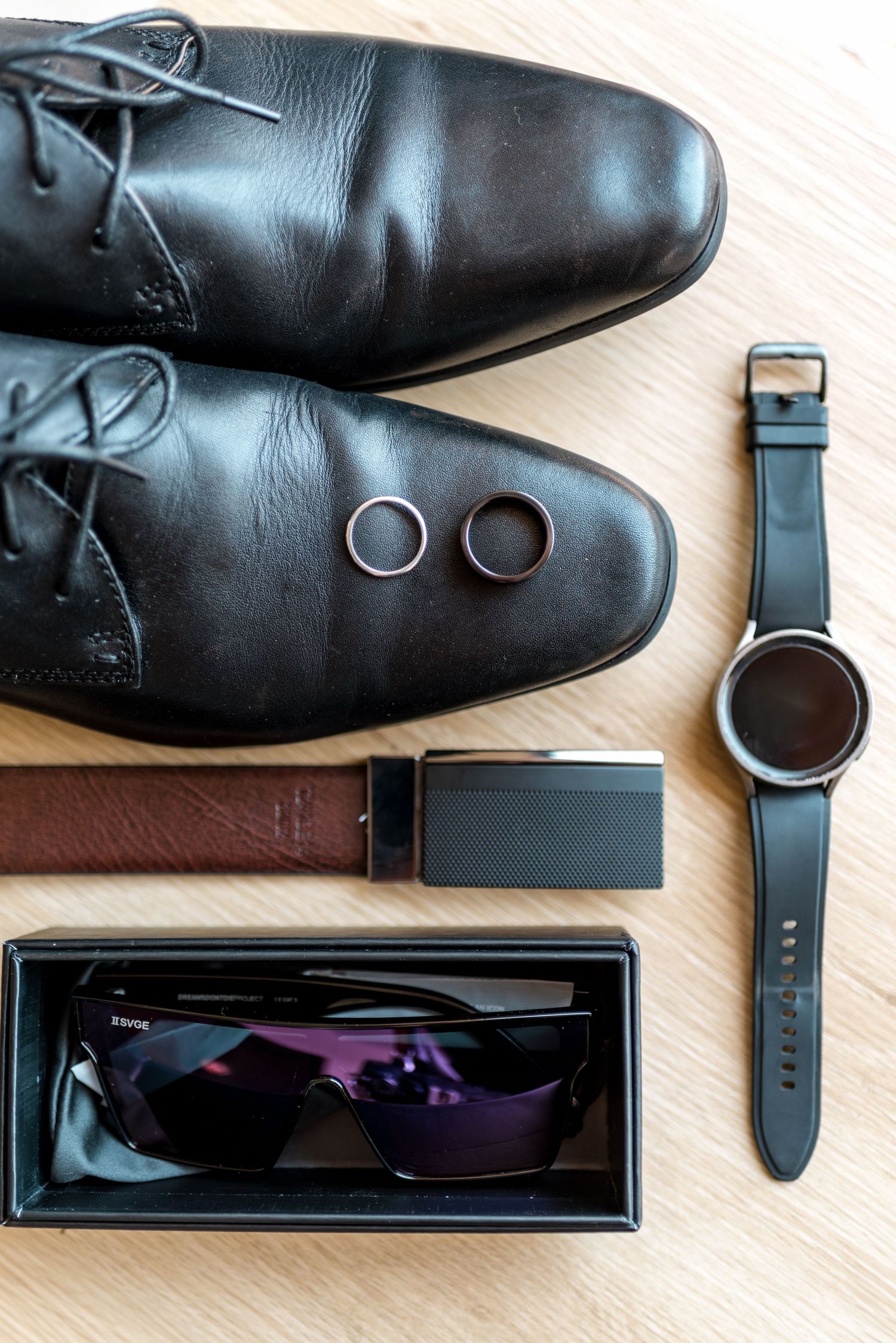 Grooms wedding flat lay featuring black dress shoes, rings, watch, belt, and sunglasses on wood.
