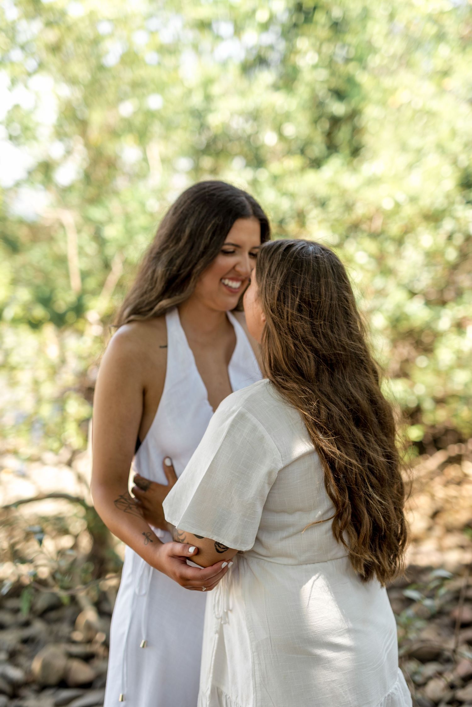 Two women in white dresses smile and embrace closely in a sun-lit outdoor setting with green trees behind them.