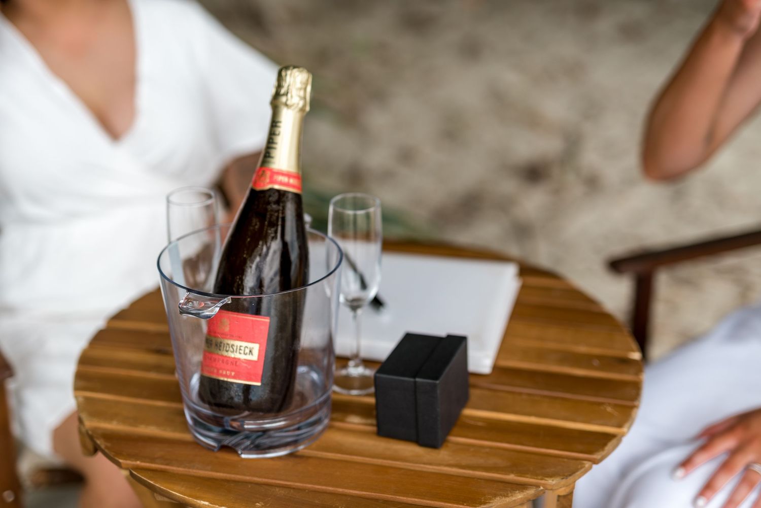 Champagne bottle in ice bucket with glasses and ring box on wooden table at beach wedding.