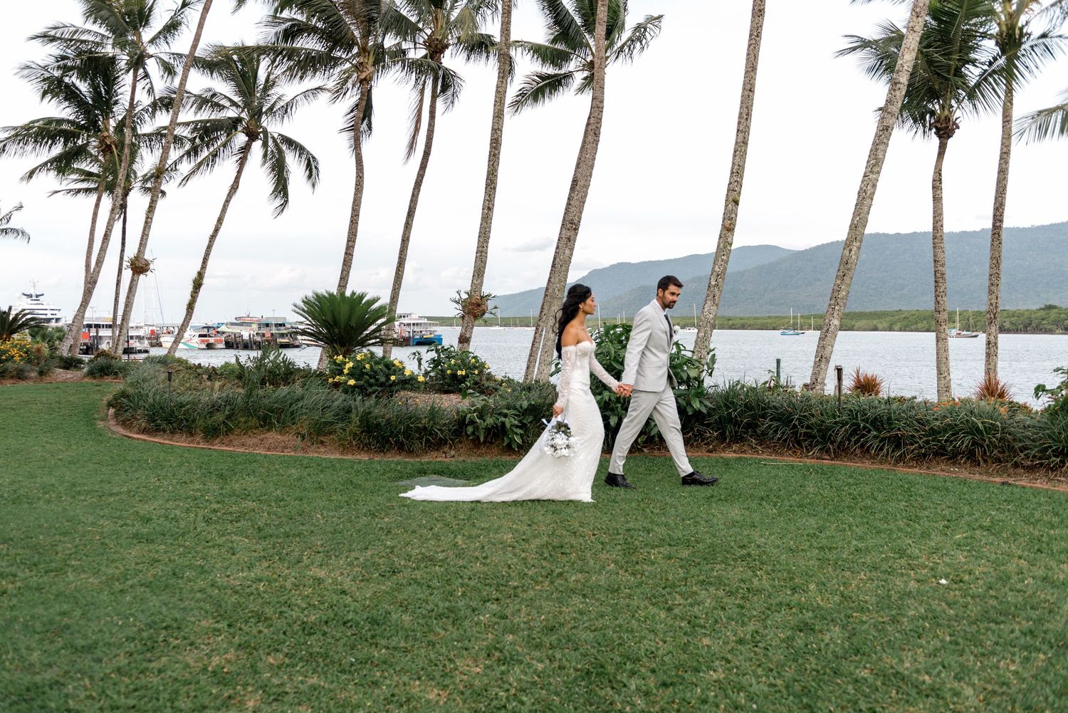Bride and groom walk along waterfront path lined with tall palm trees on their wedding day.
