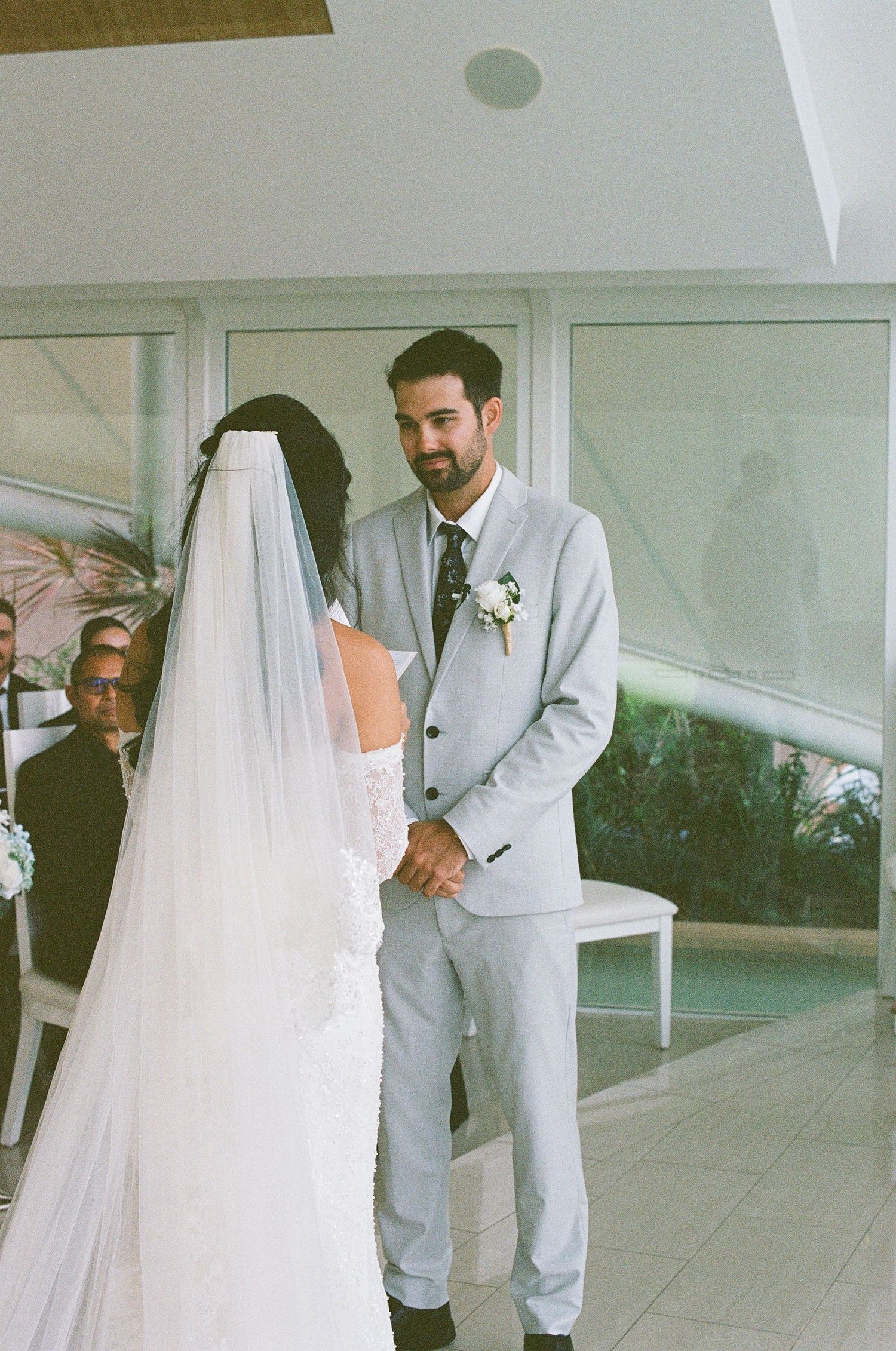 Groom in gray suit gazes lovingly at veiled bride during wedding ceremony.