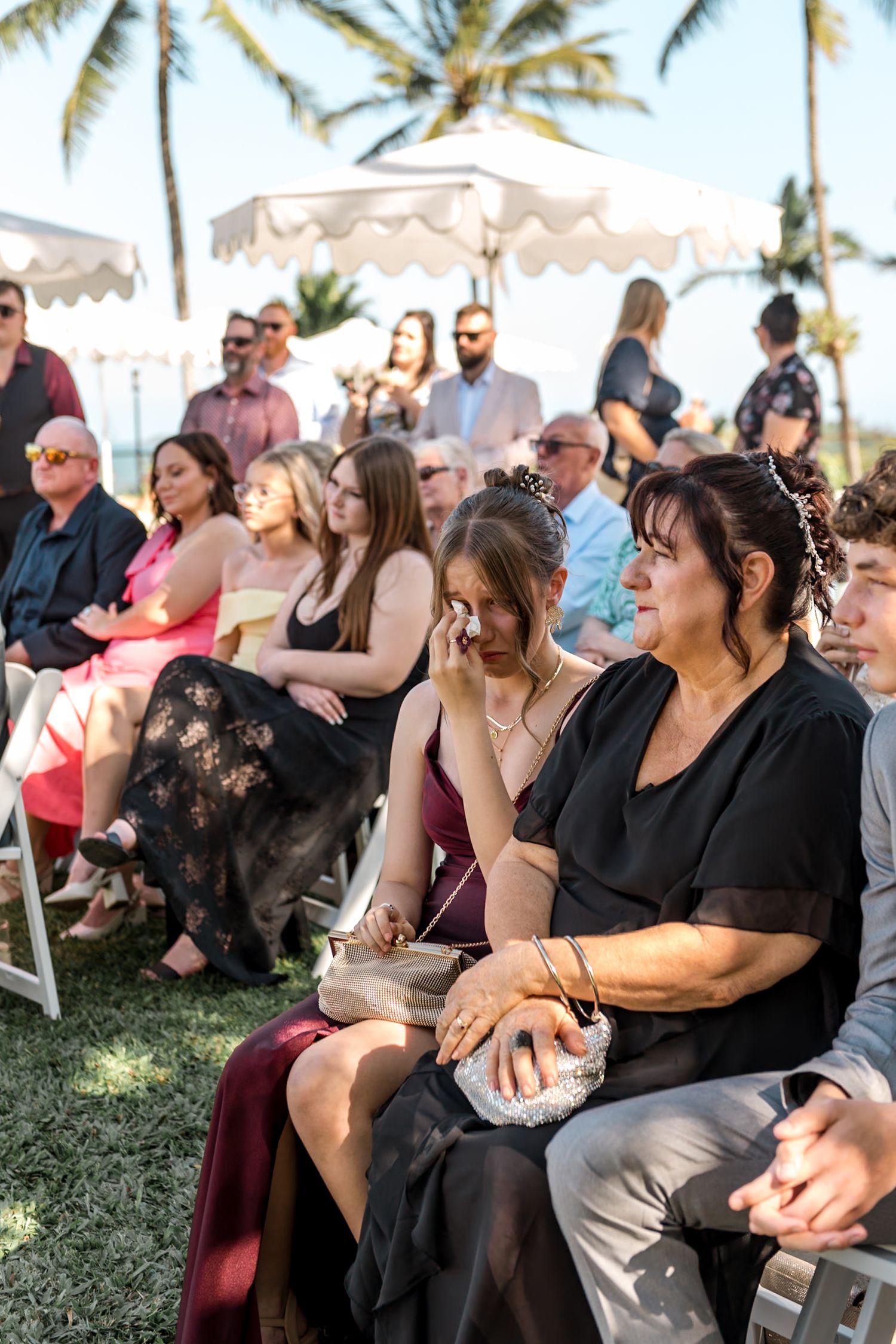 Emotional wedding guests wipe tears while watching outdoor tropical ceremony seated in white chairs on lawn.