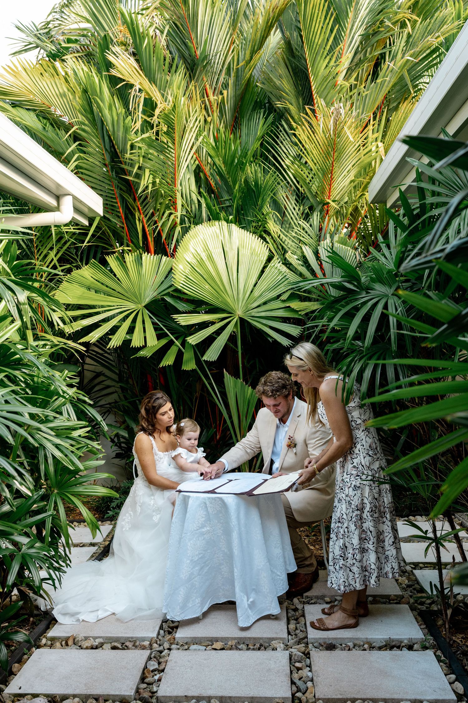 Bride, groom, and celebrant sign wedding register at outdoor table surrounded by tropical palms.