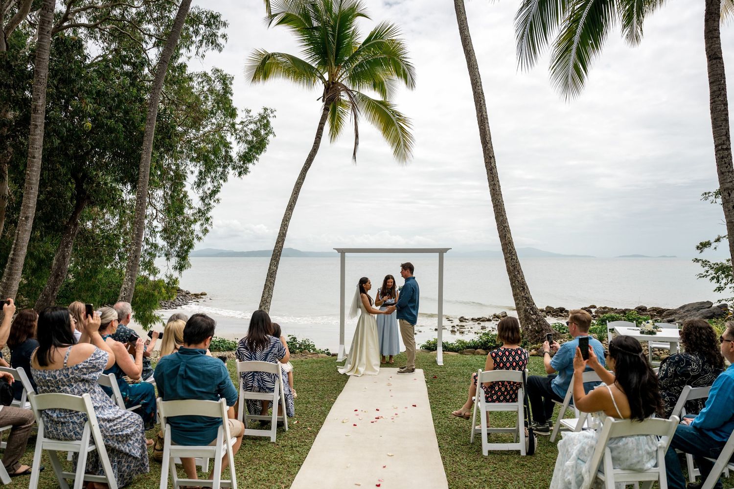 Outdoor beach wedding ceremony with guests seated on white chairs beneath palm trees by the ocean at Little Cove, Port Douglas.