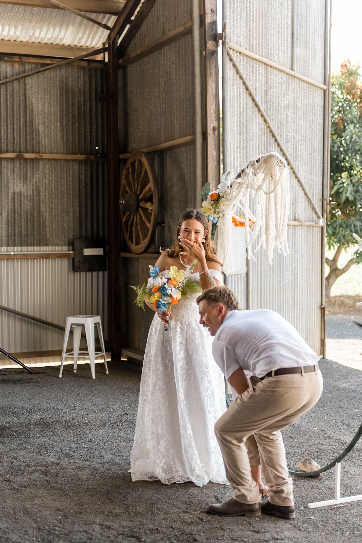 Bride in lace off-shoulder gown laughs as groom adjusts her dress train inside a rustic corrugated iron barn.