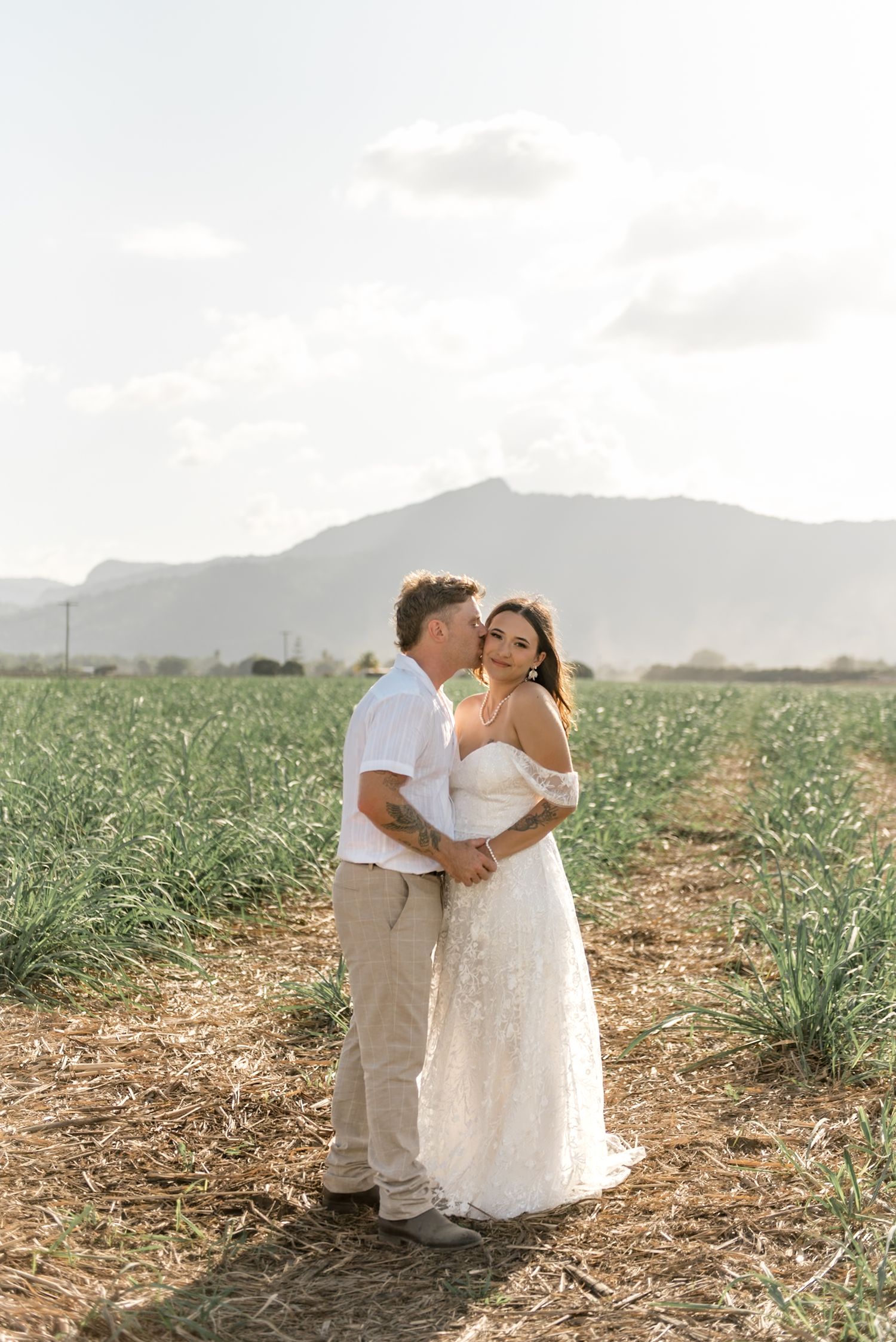 Bride and groom embracing in a lush green field with mountain backdrop on their wedding day.