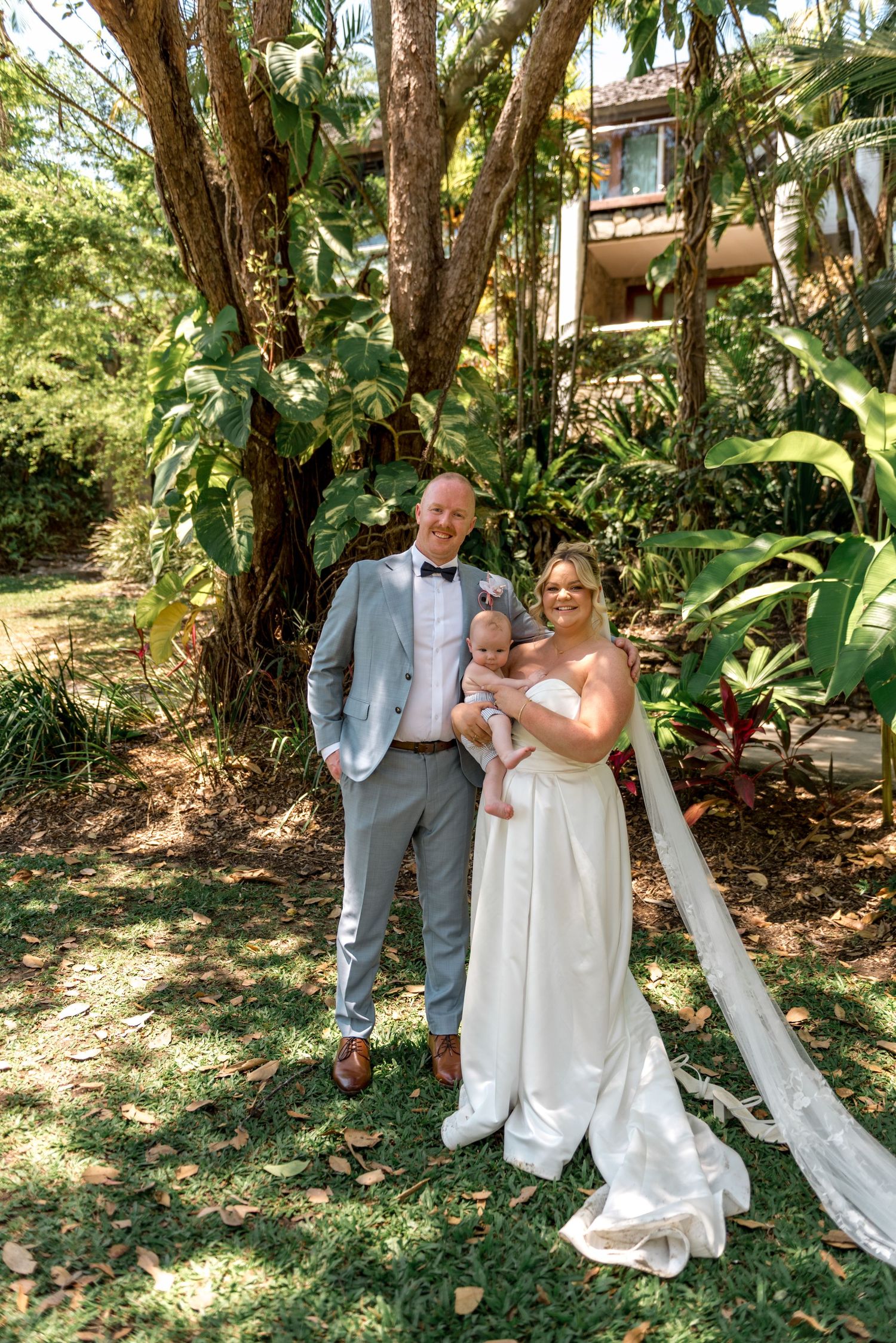 Happy newlyweds posing with their baby in a lush tropical garden on their wedding day.